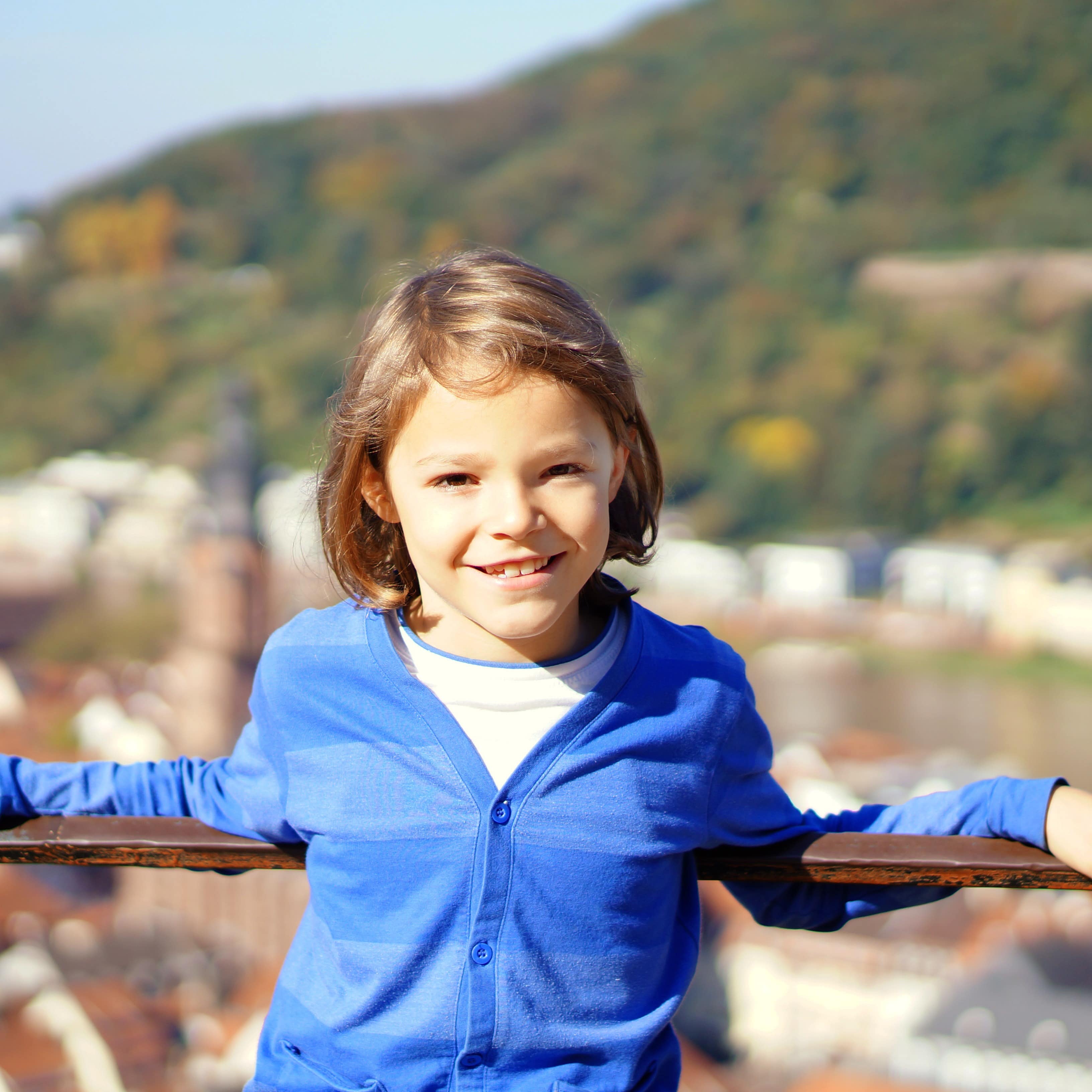 Junge mit kinnlangen Haaren und blauer Strickjacke lehnt an einem Geländer oberhalb von Heidelberg.
