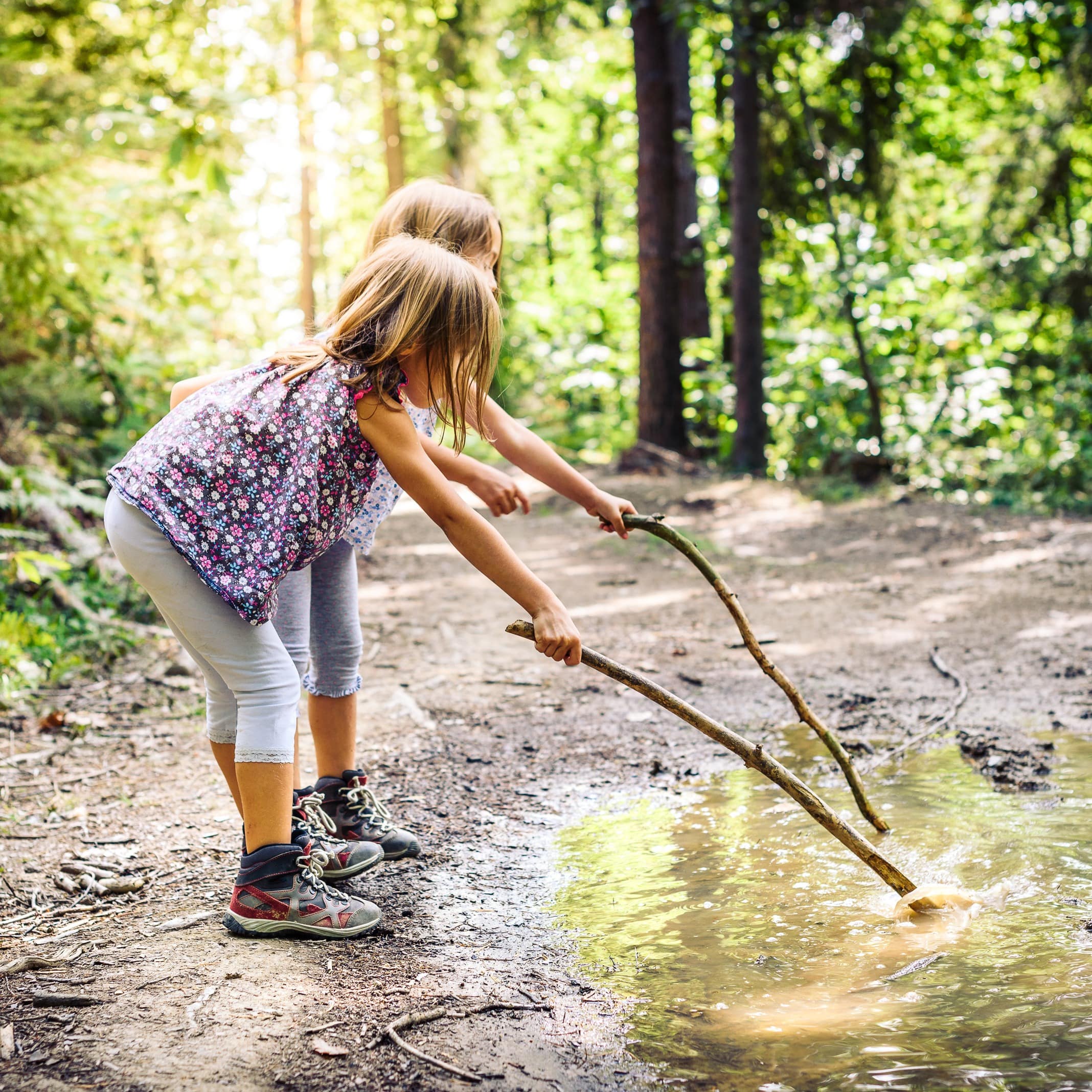 2 Mädchen mit Wanderschuhen spielen mit Stöcken im Wasser in einem Wald.