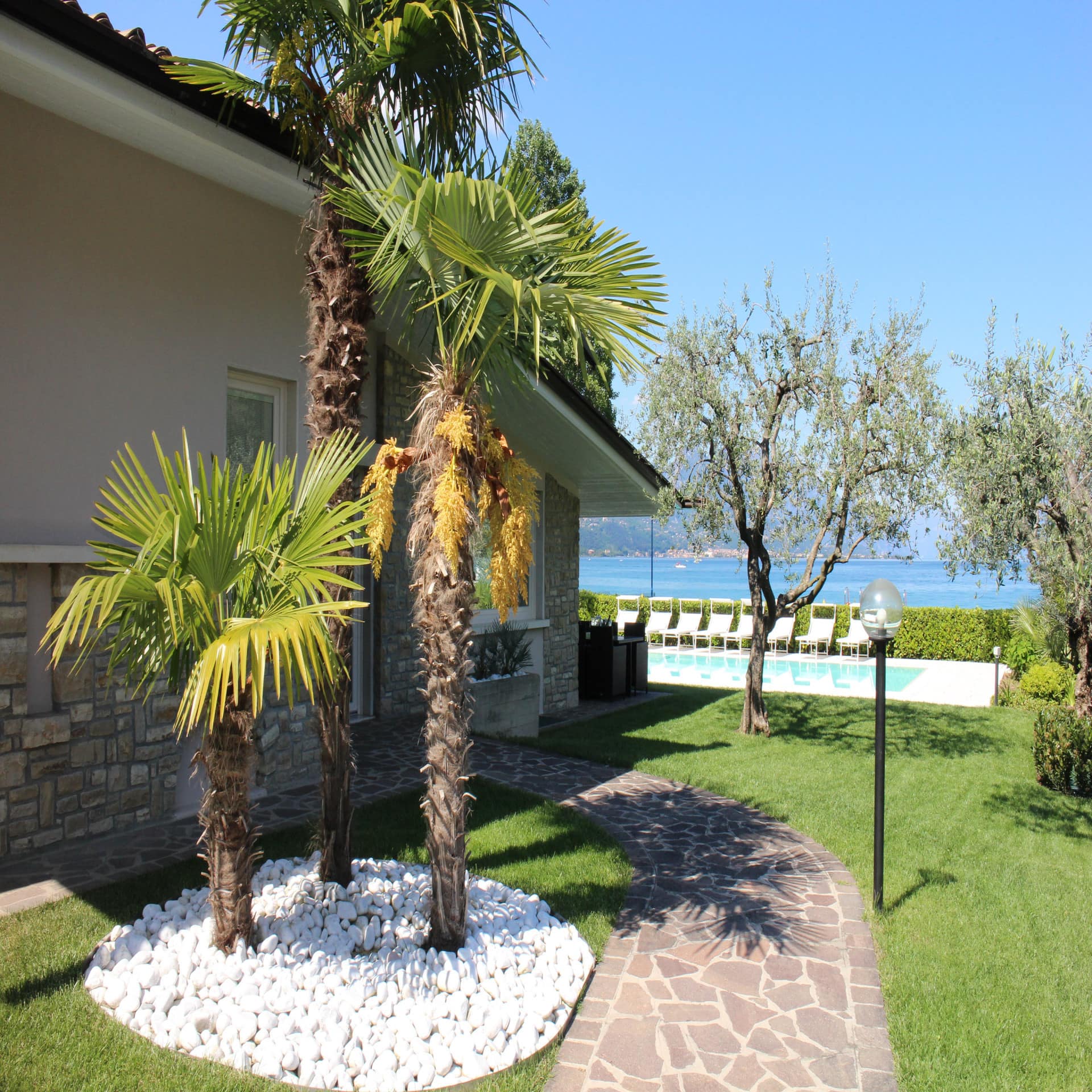 Seitlicher Blick auf ein Haus mit Palmen, Garten und Pool mit Sonnenliegen direkt am Gardasee.