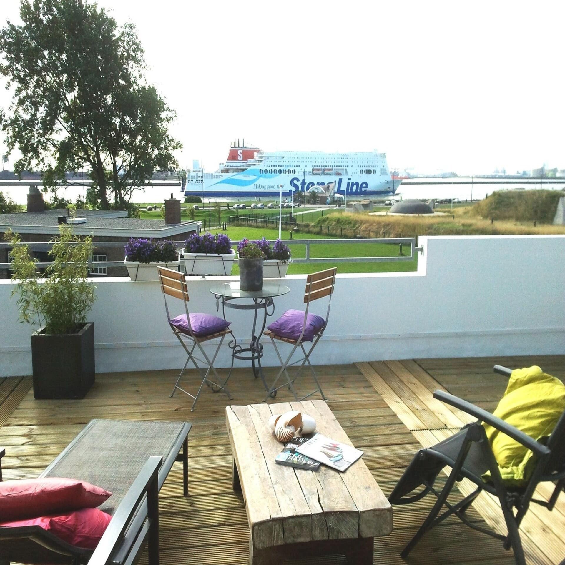 Großer Balkon mit Liegestühlen, Tisch und Stühlen mit Blick über eine Wiese auf das Meer und ein Schiff.