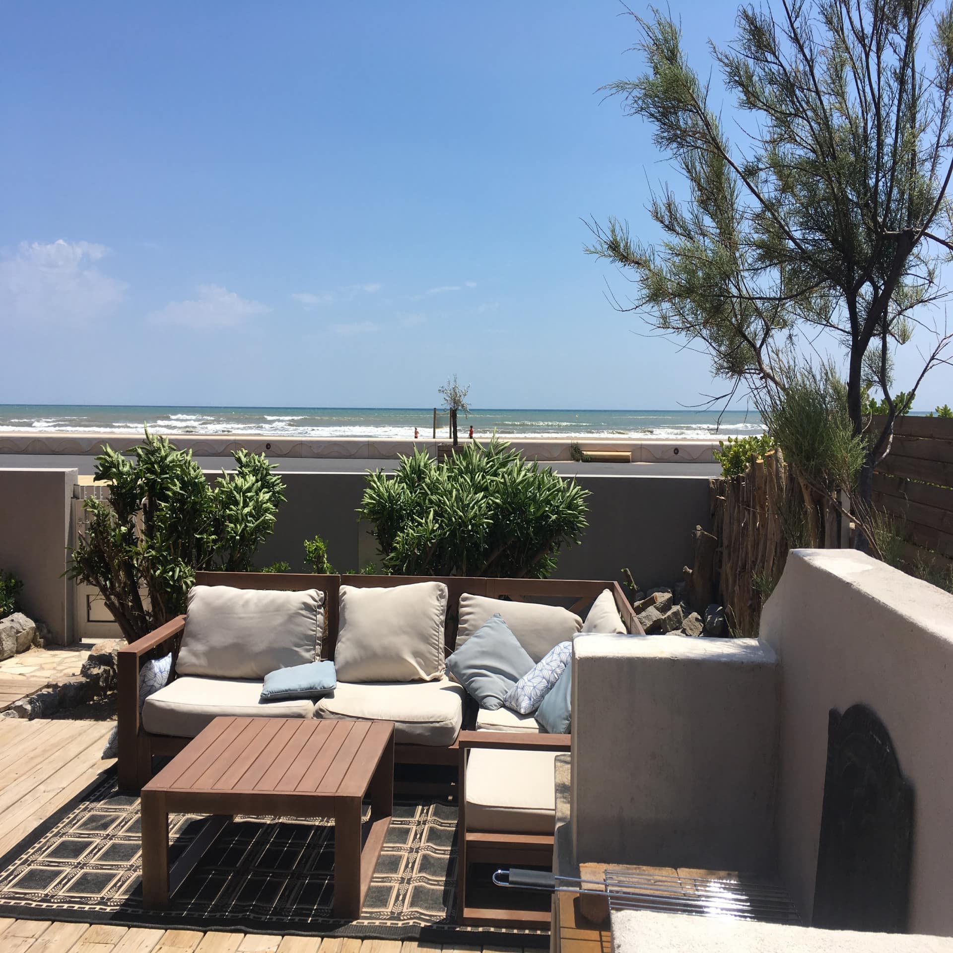 Terrasse mit Sofa, Tisch und Grill sowie Blick auf das Meer. Die Sonne scheint.