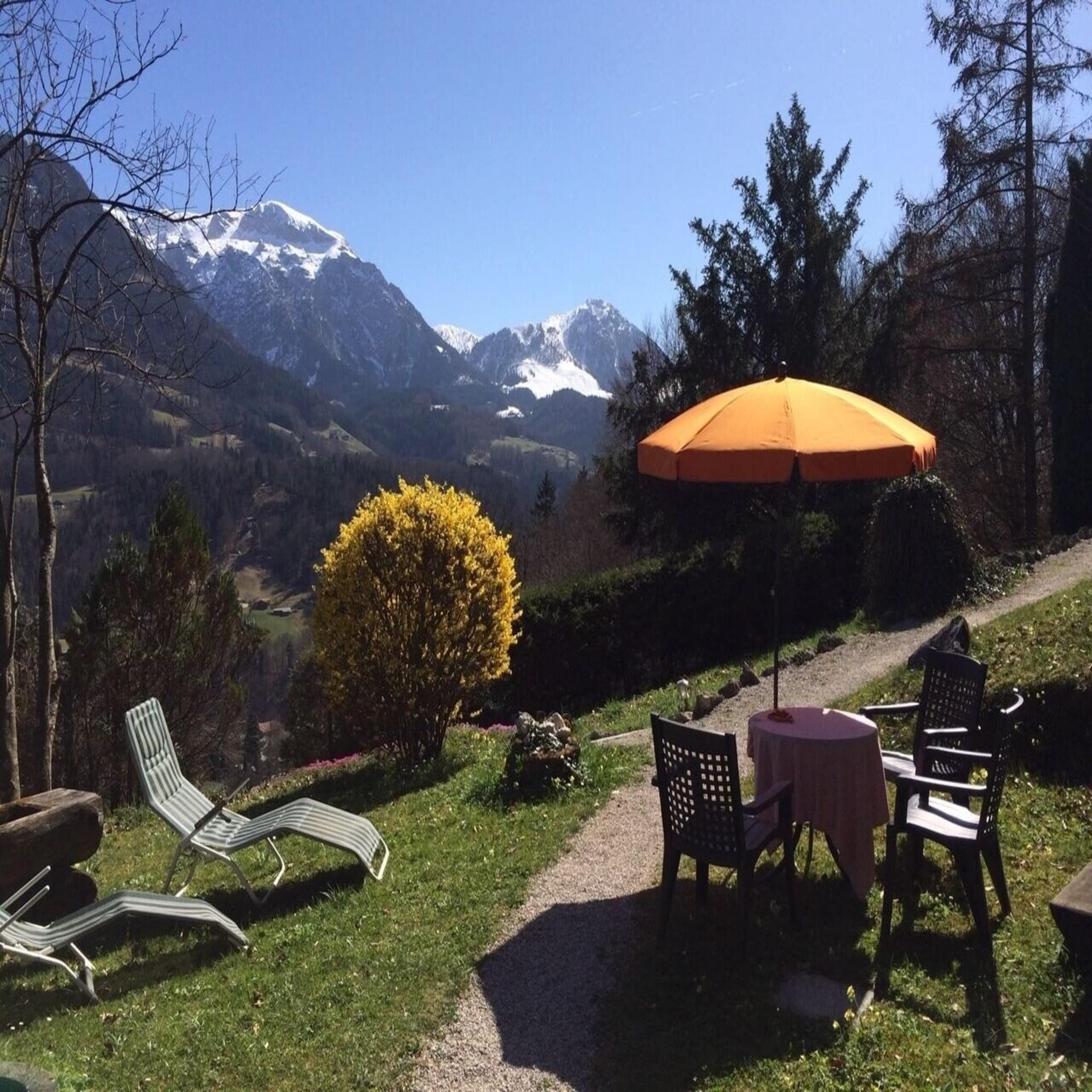 Berggarten mit Tisch, Sonnenschirm und Sonnenliegen und Blick auf die Berge. 