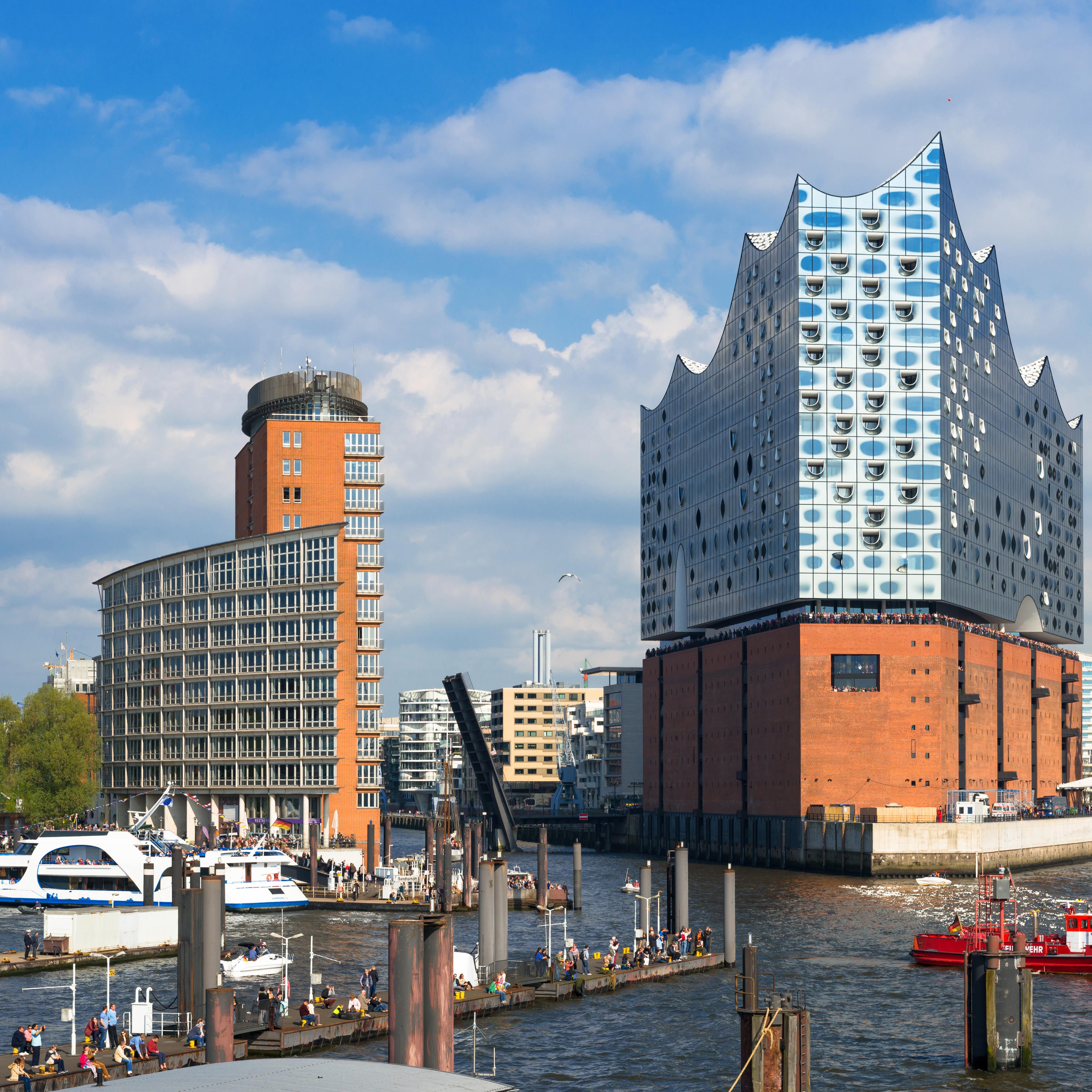 Blick auf de HafenCity und die Elbphilharmonie im Sonnenschein. Auf einem Steg sind Menschen, im Wasser Boote.