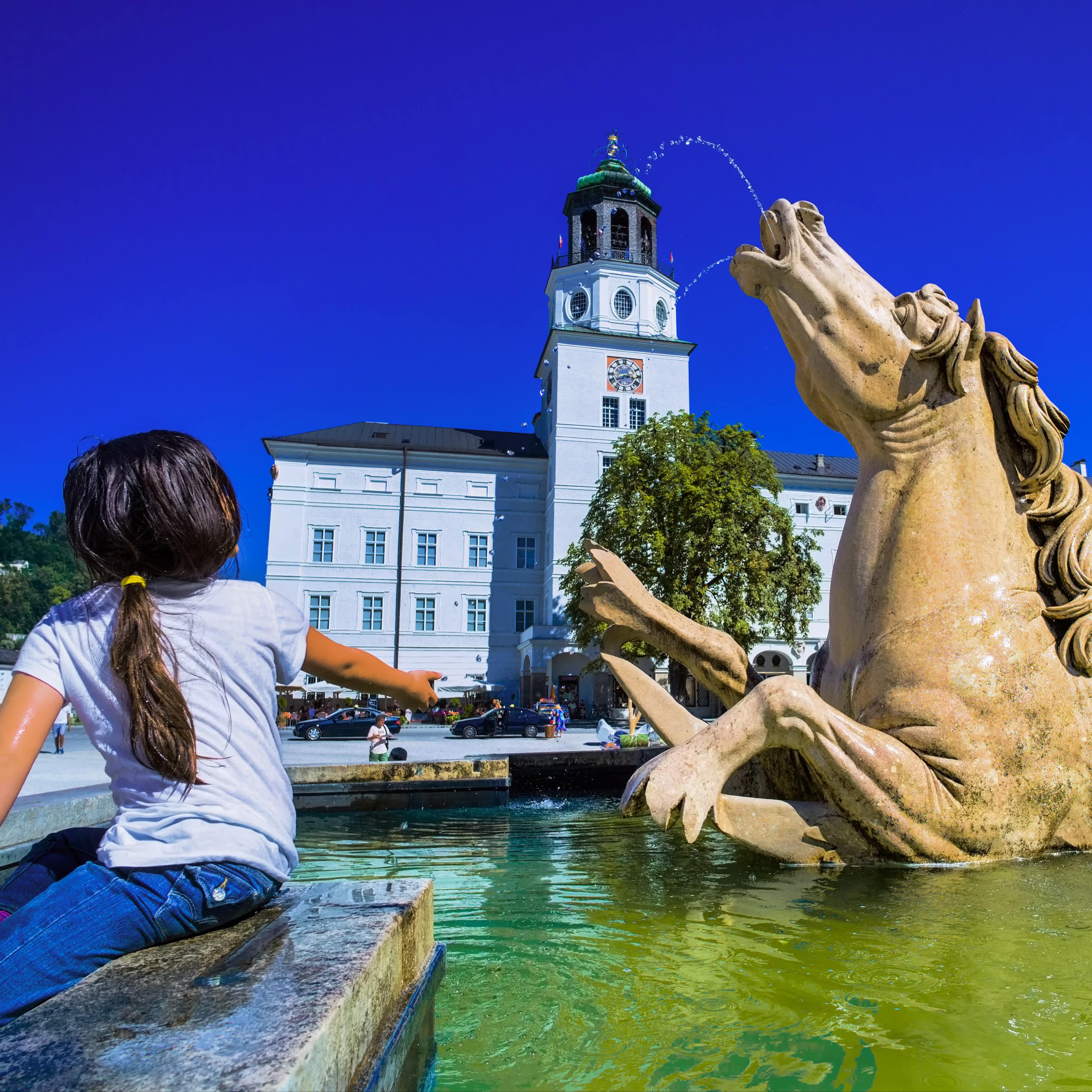 Rückenansicht: Ein Mädchen am Brunnen auf dem Residenzplatz in Salzburg. Image URL: