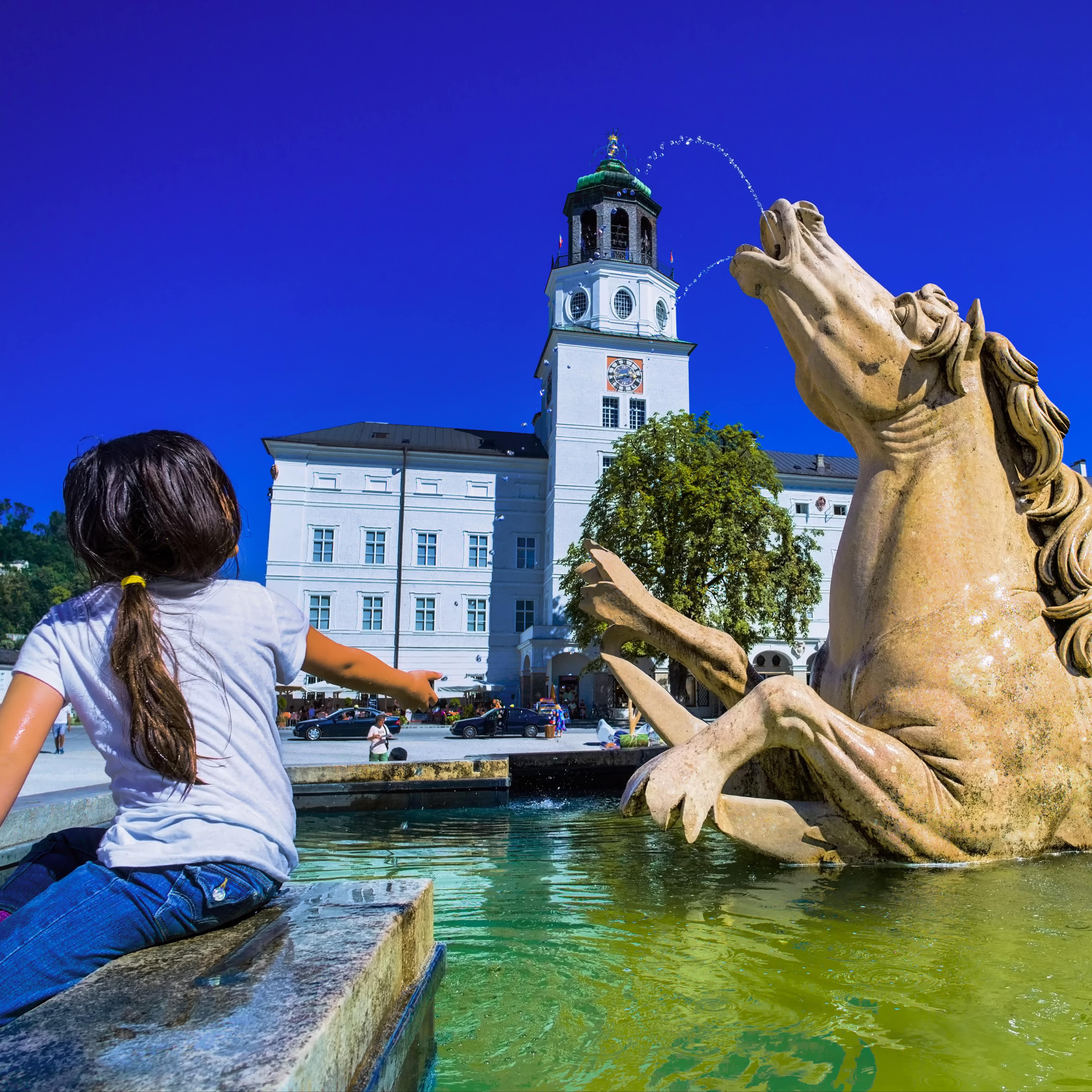 Rückenansicht: Ein Mädchen am Brunnen auf dem Residenzplatz in Salzburg. Image URL: 