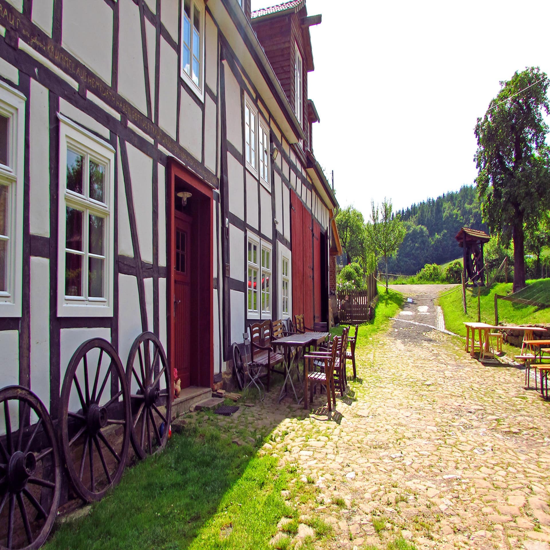 Innenhof eines Fachwerk-Bauernhauses mit einem großen Tisch davor. Wagenräder lehnen an der Wand.