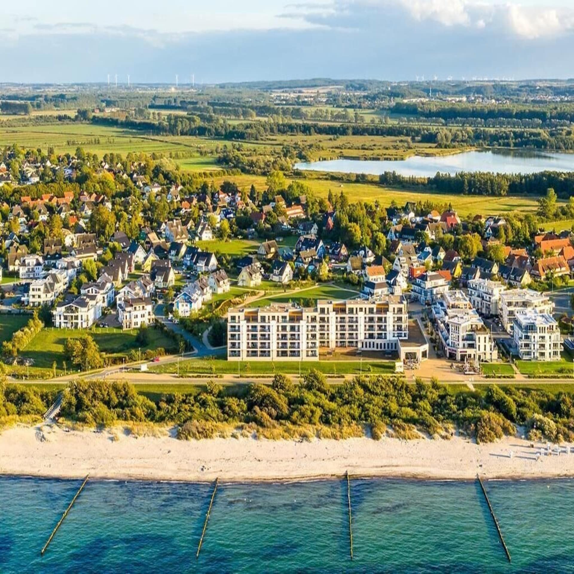 Blick aus der Luft auf einen Ferienhauspark direkt an der Ostsee. Im Hintergrund ein See und Grünes. 