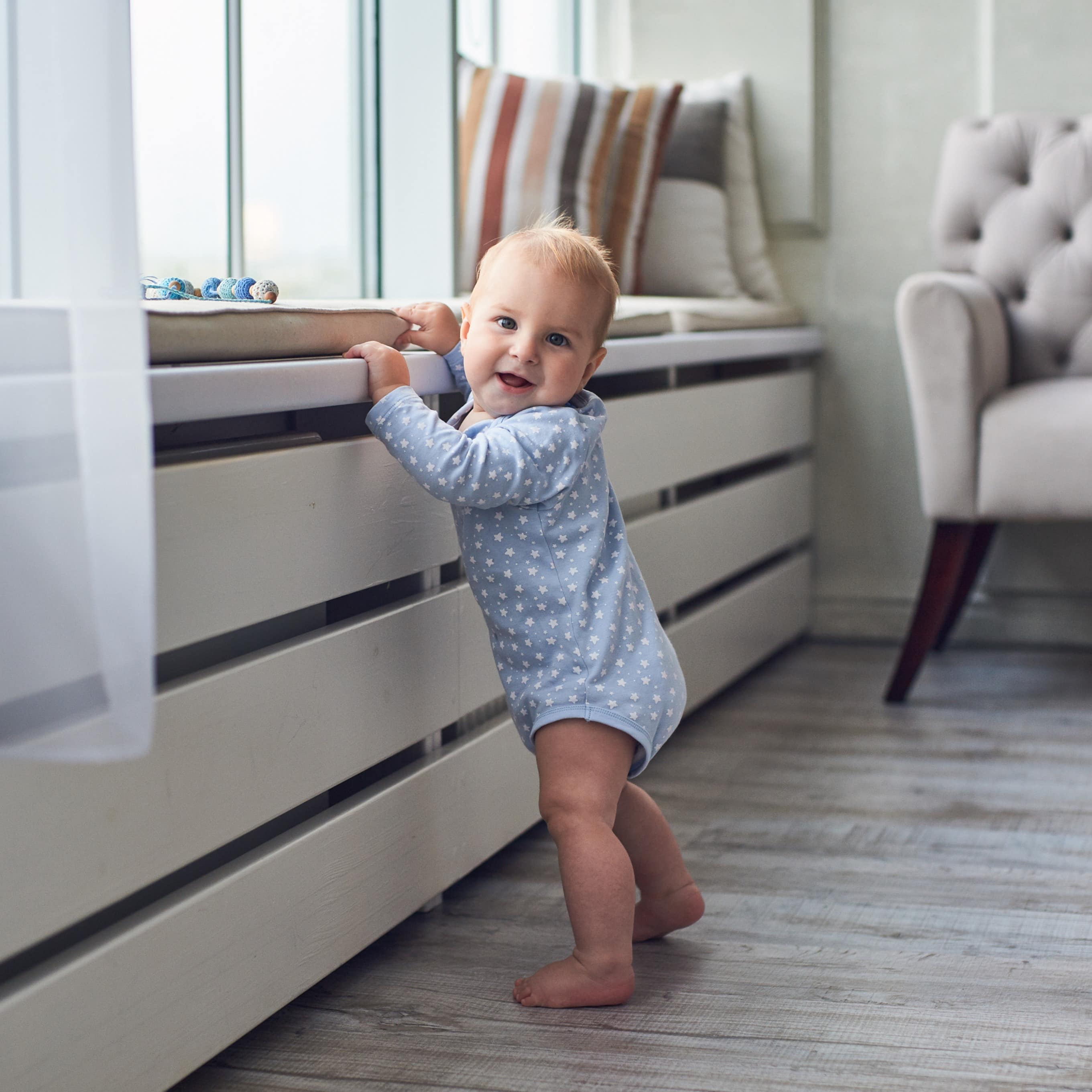 Baby im Strampler steht und hält sich an einer Fensterbank fest. Es schaut in die Kamera. 