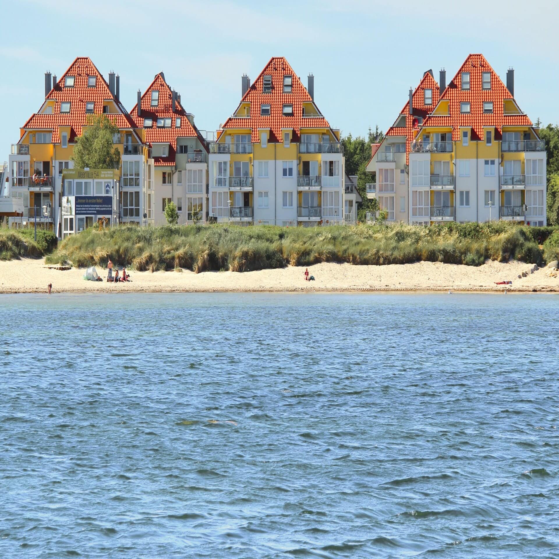 Blick vom Wasser auf den Strand an dem Leute sitzen, dahinter Mehrfamilienhäuser. Die Sonne scheint.